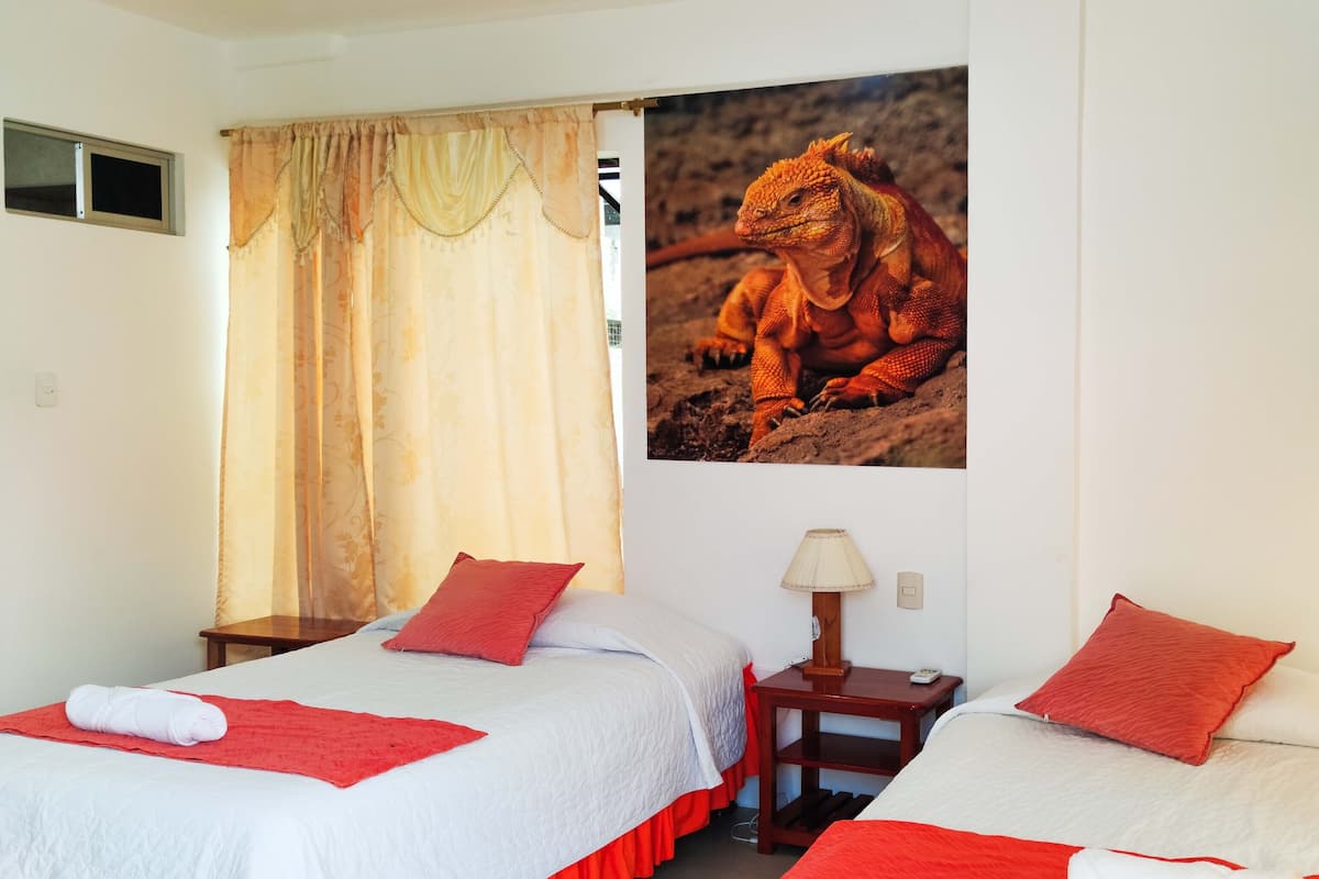Une chambre d’hôtel avec deux lits, chacun recouvert d’une courtepointe blanche et orange, agrémentée d’oreillers rouges et d’une serviette roulée. On y trouve une fenêtre avec un rideau, une lampe fixée au mur et un tableau représentant un lézard accroché au mur.