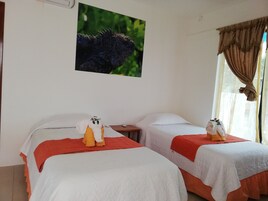 Habitación de hotel con dos camas, cada una con una colcha blanca y una manta roja. Hay una mesita con un control remoto y un marco de fotos en la pared sobre las camas.