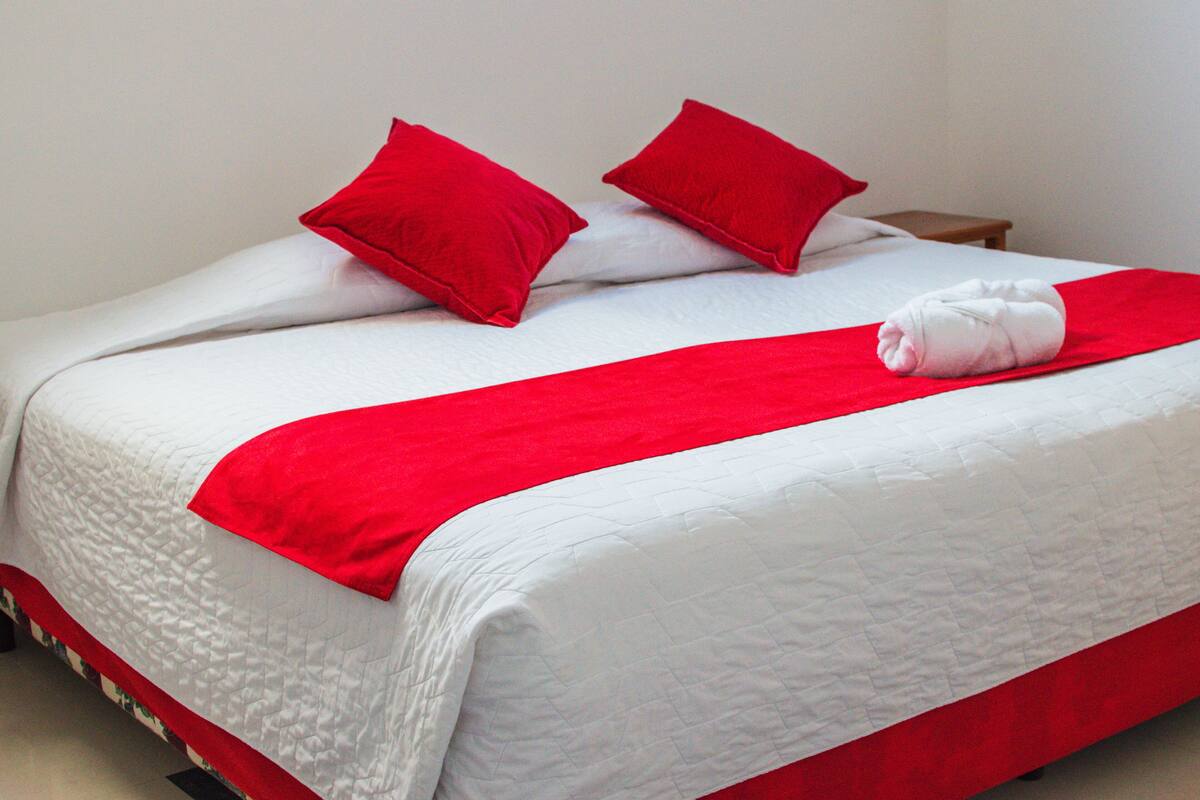 Un lit bien fait, recouvert d’une couverture rouge et blanche, avec deux oreillers rouges et une serviette blanche.