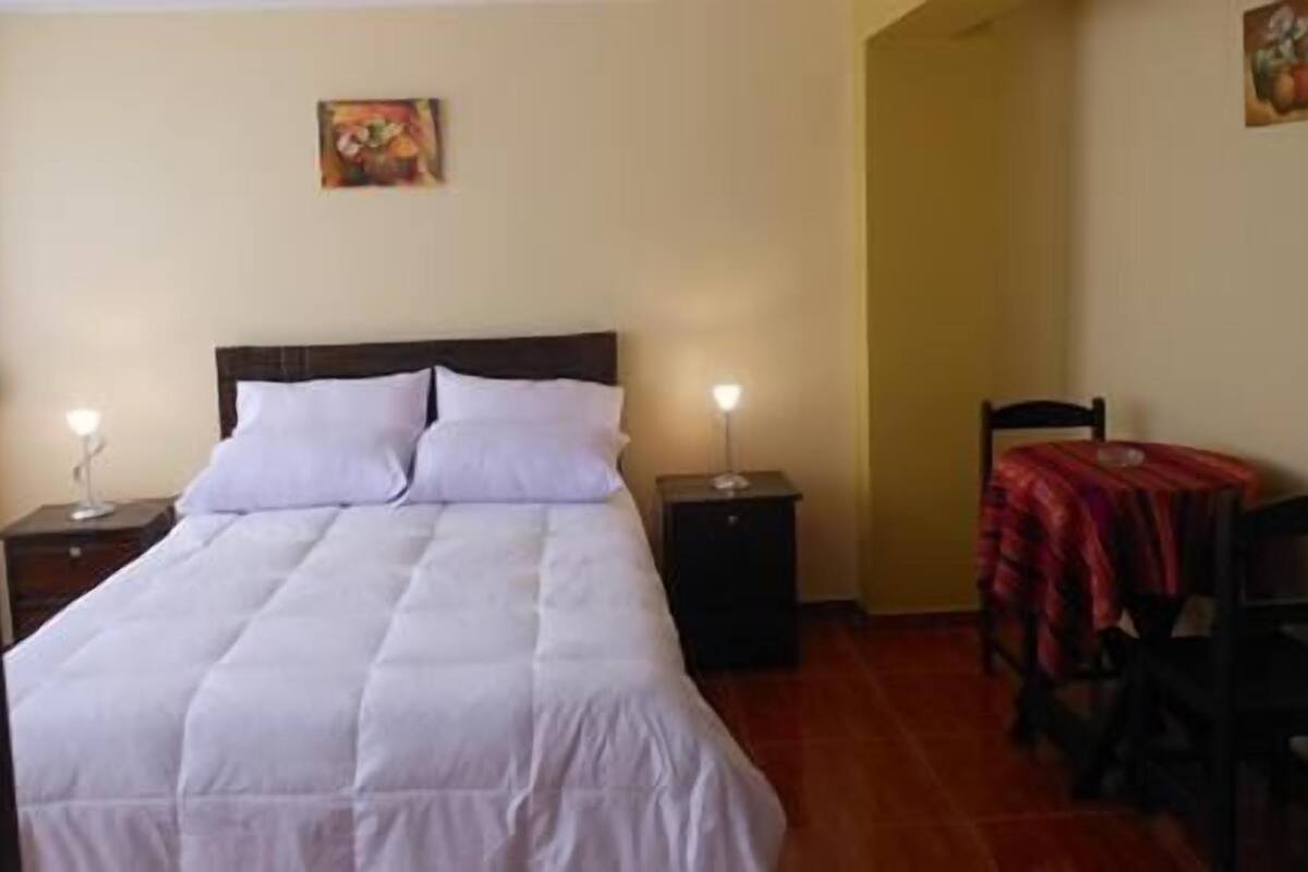 A neatly made bed with white linens, two bedside tables with lamps, a small table with a red tablecloth, and a chair.