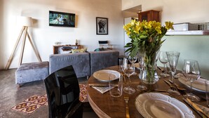 Un comedor con una mesa redonda preparada para seis personas, un sofá, un televisor y un jarrón con flores amarillas.