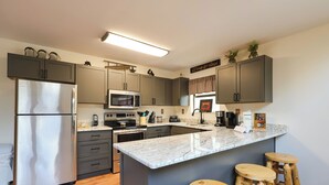 A modern kitchen with a stainless steel refrigerator, a microwave, a stove, and a sink. There are wooden stools and a marble countertop.