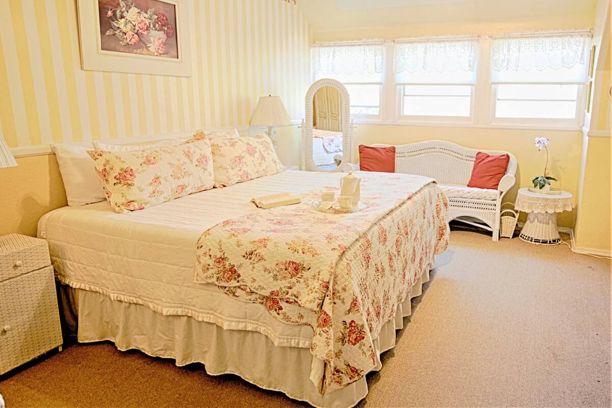 A bedroom with a large bed, a bedside table, a chair, a small table with a lamp, and a window with curtains.