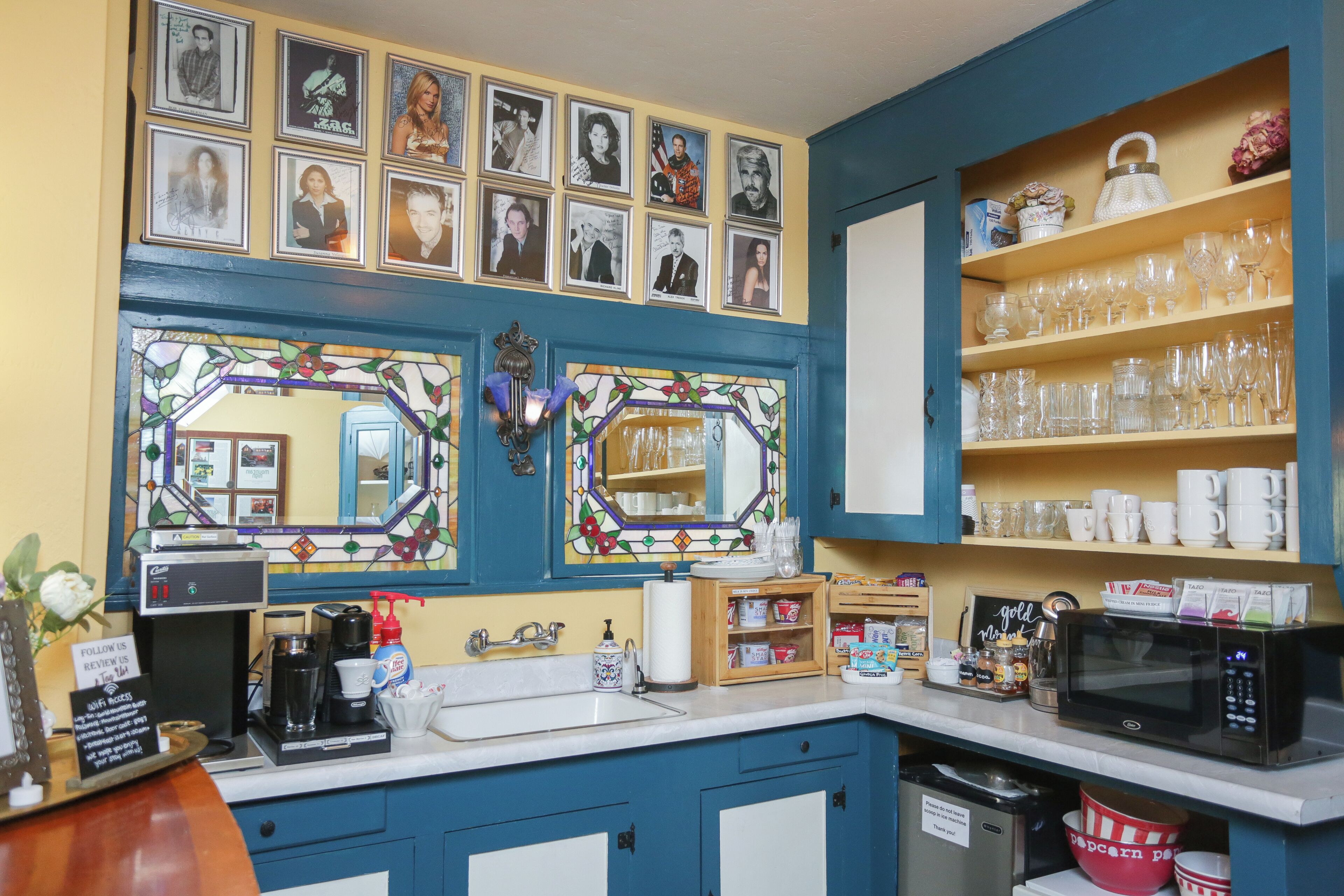 Una cocina con armarios azules, una cafetera y un microondas. Hay cuadros en la pared y estantes con vasos y tazas.
