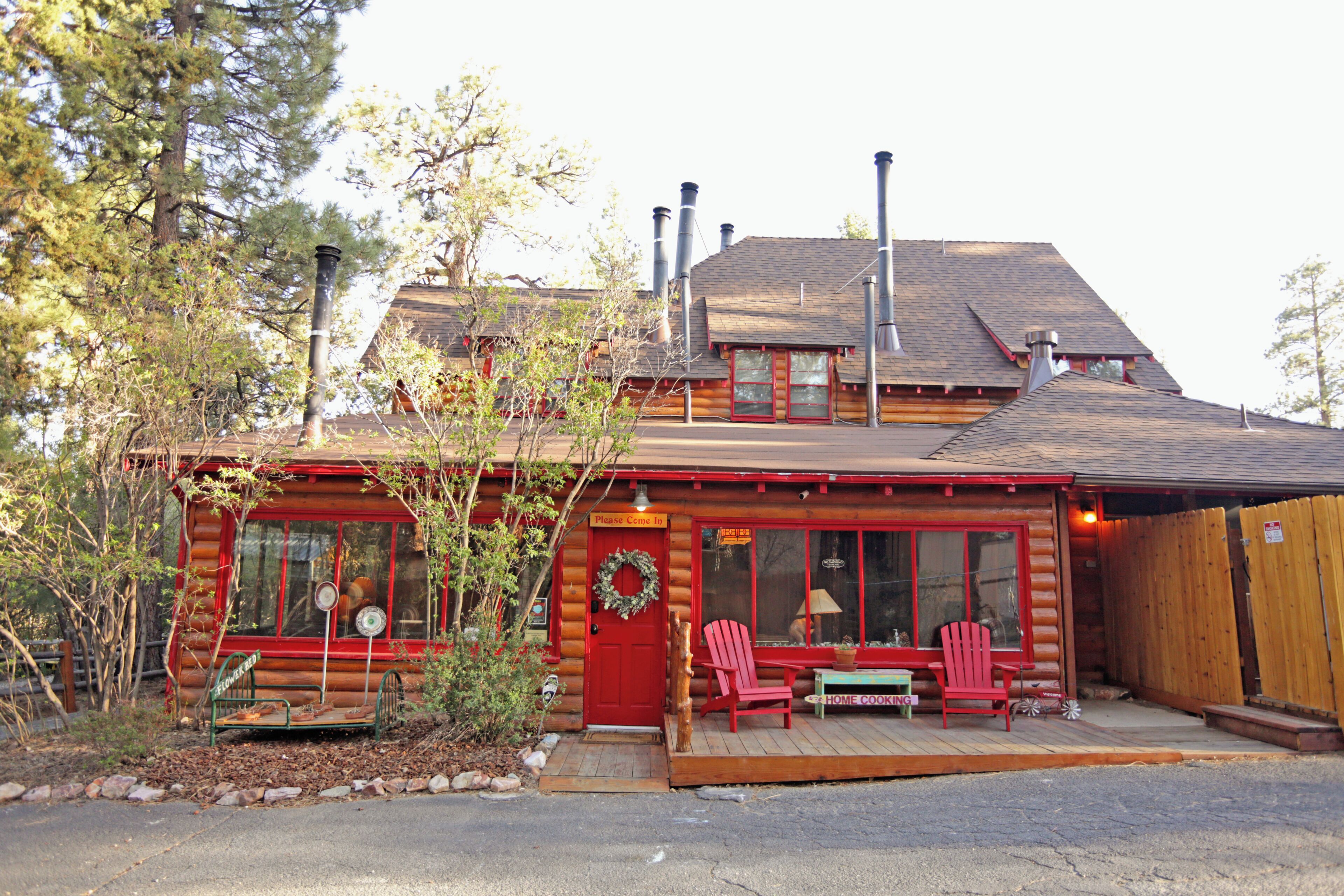 A log cabin with a red door, two windows, a porch with red Adirondack chairs, and a sign that reads 'Please Come In.'