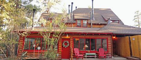 A log cabin with a red door, two windows, a porch with red Adirondack chairs, and a sign that reads 'Please Come In.'