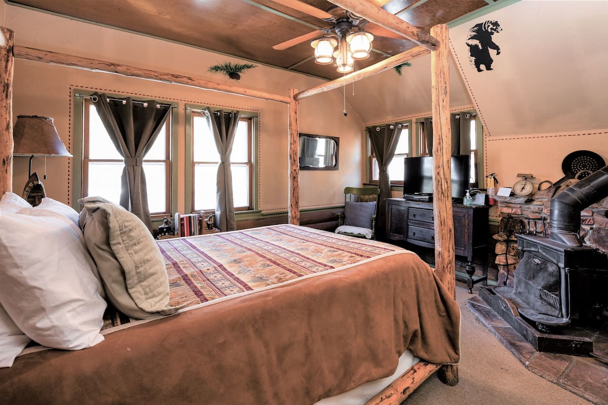 A rustic bedroom with a four-poster bed, a wooden ceiling, a fireplace, and a television on the wall.