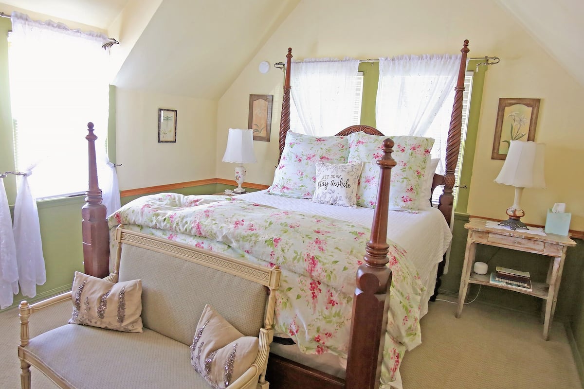 A four-poster bed with floral bedding, a bedside table with a lamp, and a chair with decorative pillows.