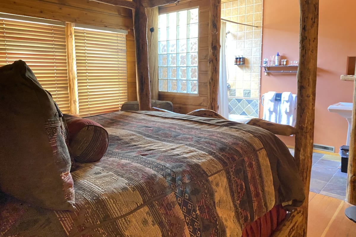 A cozy room with a large bed, wooden beams, a window with blinds, and a view of the outside balcony.