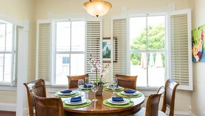 A dining room with a round table set for four, white shutters on the windows, and a painting on the wall.
