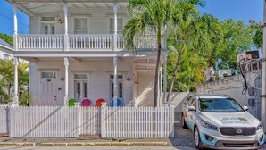 A two-story white building with a balcony, a white picket fence, and a Kia vehicle parked in front.