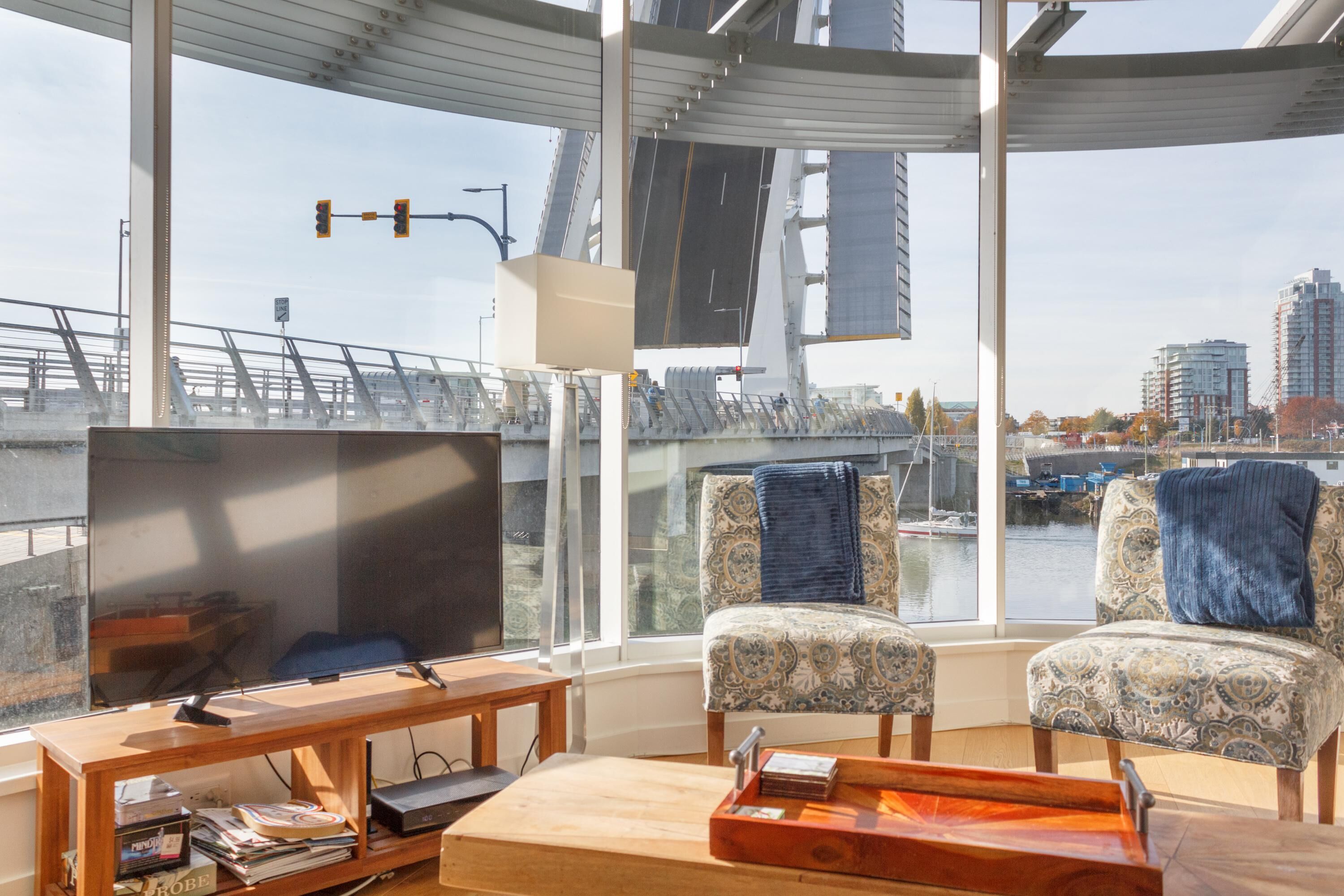 Una sala moderna con un televisor grande, dos sillones estampados, una mesa de centro de madera y vistas a un paisaje urbano y un puente a través de ventanas amplias.