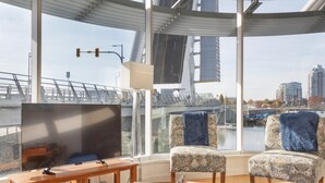 Un salon moderne doté d’un grand téléviseur, de deux fauteuils à motifs, d’une table basse en bois et offrant une vue sur un paysage urbain et un pont, le tout visible à travers de grandes fenêtres.