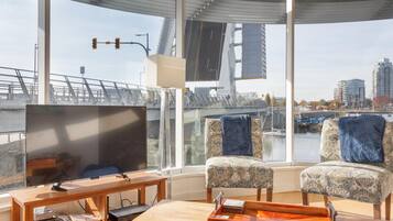 Una sala moderna con un televisor grande, dos sillones estampados, una mesa de centro de madera y vistas a un paisaje urbano y un puente a través de ventanas amplias.