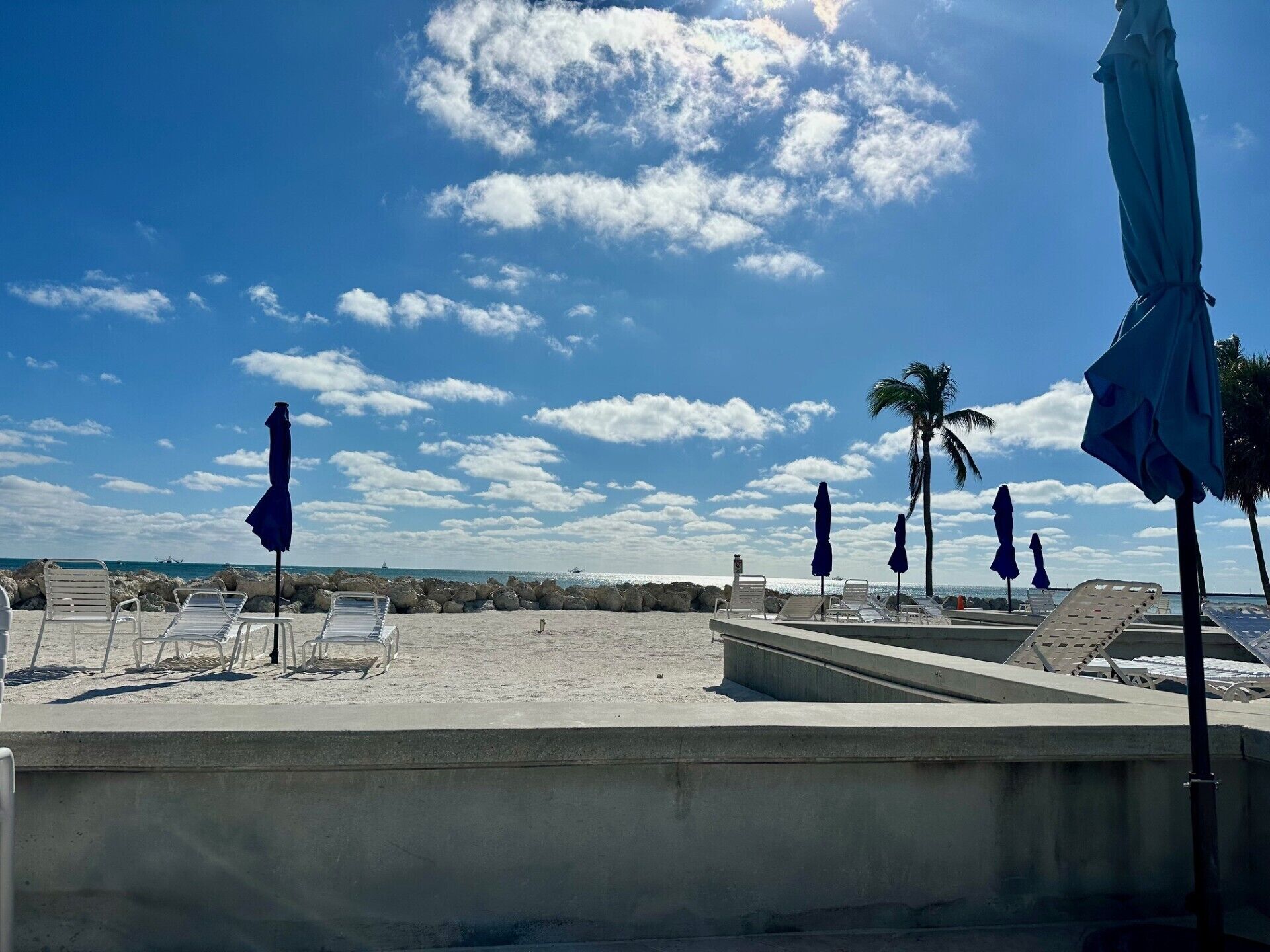 A beach with lounge chairs and umbrellas, a concrete pool, and a view of the ocean and sky.