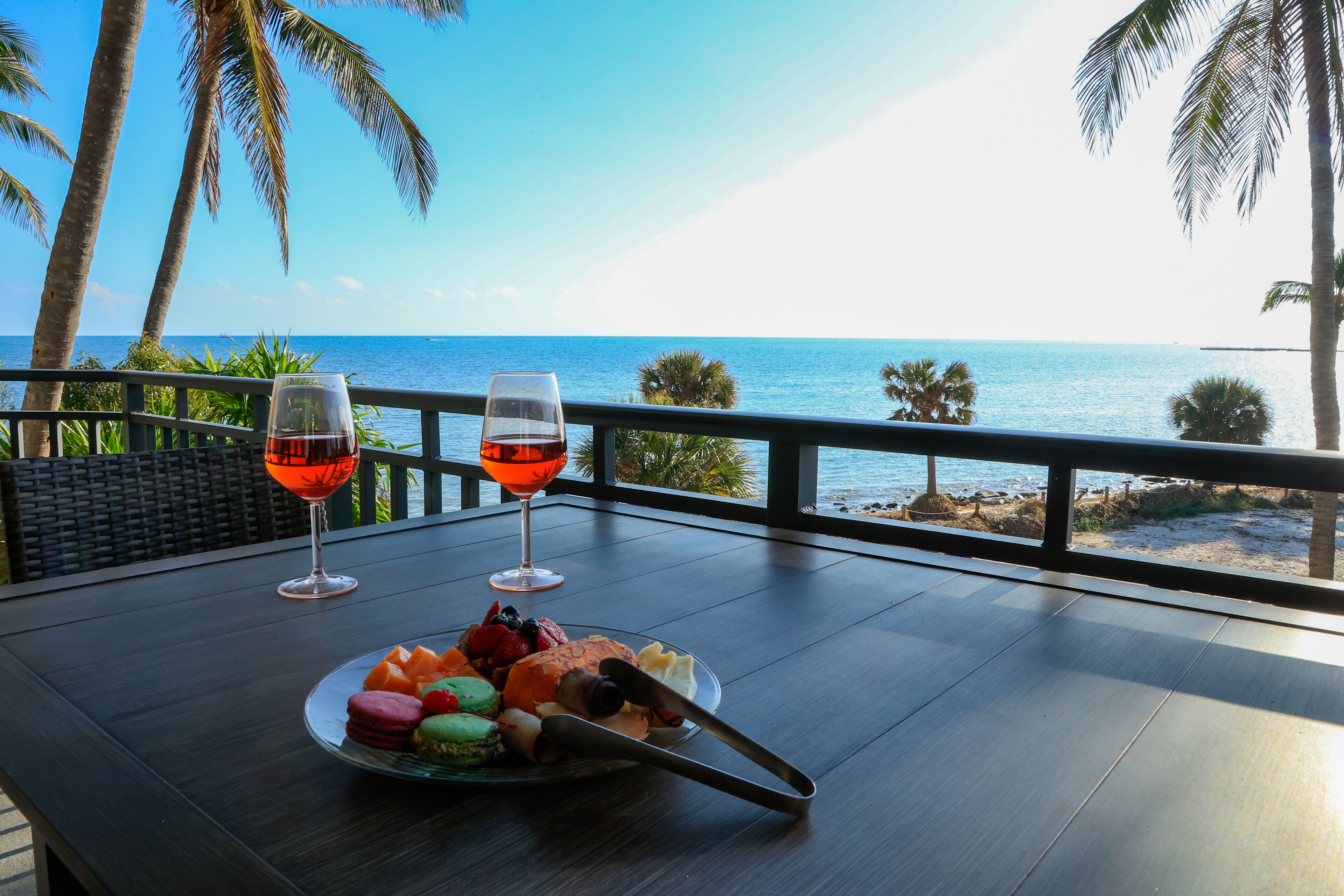 A table with a plate of food and two glasses of red wine, overlooking a beach with palm trees.