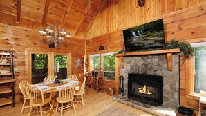 Interior de una cabaña de madera con chimenea de piedra, una mesa de comedor, sillas y un televisor colocado sobre la chimenea.