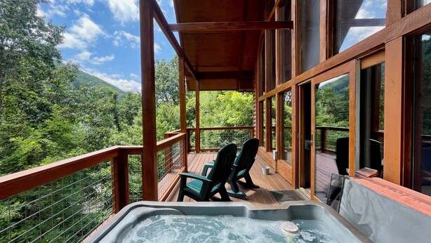 Une terrasse en bois avec un jacuzzi, des chaises et une vue sur une zone montagneuse boisée.