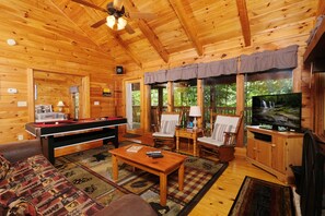 A wooden cabin interior with a pool table, comfortable seating, a television, and large windows.