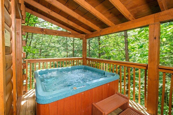 A wooden deck with a hot tub and a bench, surrounded by a forest.