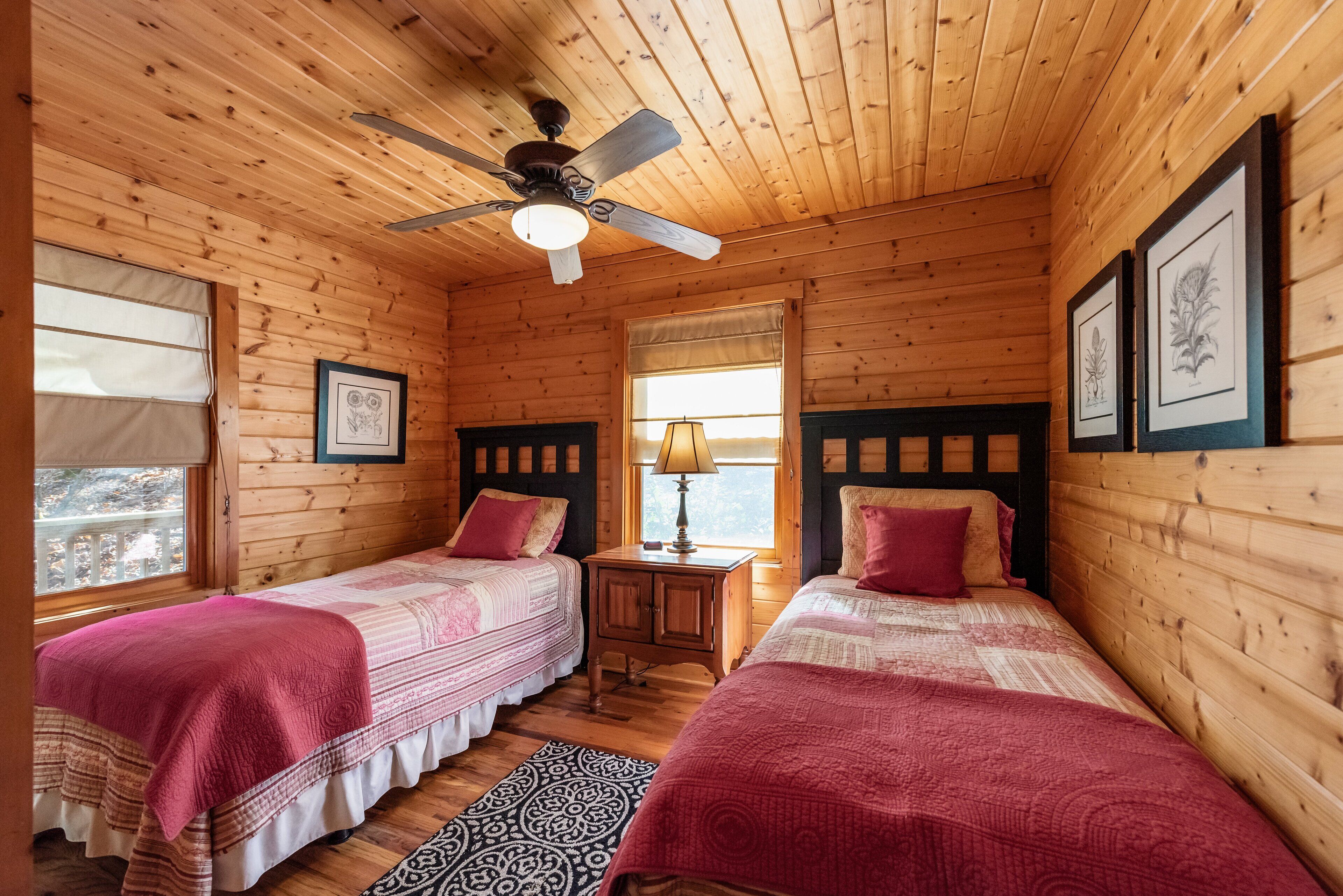 A cabin-style room with two beds, a ceiling fan, a window with a view, and framed artwork on the walls.