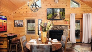 A wooden cabin interior with a dining table set for four, a fireplace, a mounted TV, and a game table. - Outdoor TV on screened in porch, hot tub, arcade game, ticket to Dollywood (Sevierville)