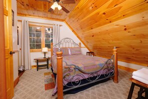 Un dormitorio con techo de madera, cama con estructura metálica, mesita de noche, lámpara y ventana con cortinas.