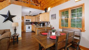 A spacious living area with a wooden dining table set for four, a kitchen with wooden cabinets, and a large window with a view of greenery.