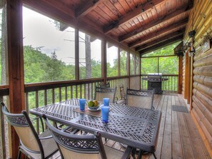 A covered outdoor deck with a table and chairs, a grill, and a view of the forest. - 6BR close to PF Parkway- 2 hot tubs, game room, 4 decks, 3 living areas! (Pigeon Forge)