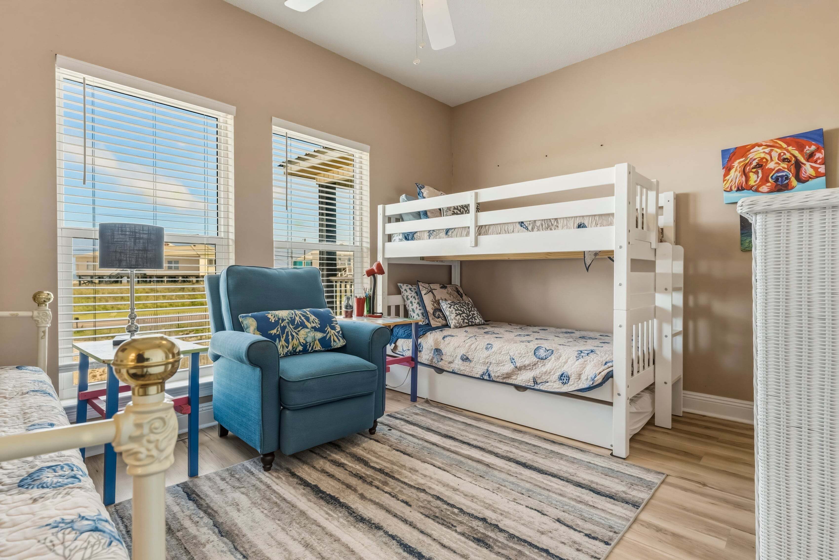 Une chambre avec un lit superposé, un fauteuil bleu, une petite table et une fenêtre avec des stores.