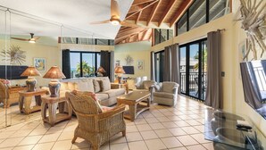 Amplia sala de estar con mobiliario de mimbre, ventilador de techo, ventanas grandes y vistas al exterior.