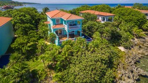 A turquoise two-story house with a red roof, surrounded by lush greenery and a clear view of the ocean. - West End waterfront w/Amenities and Views to Match! (Roatan)