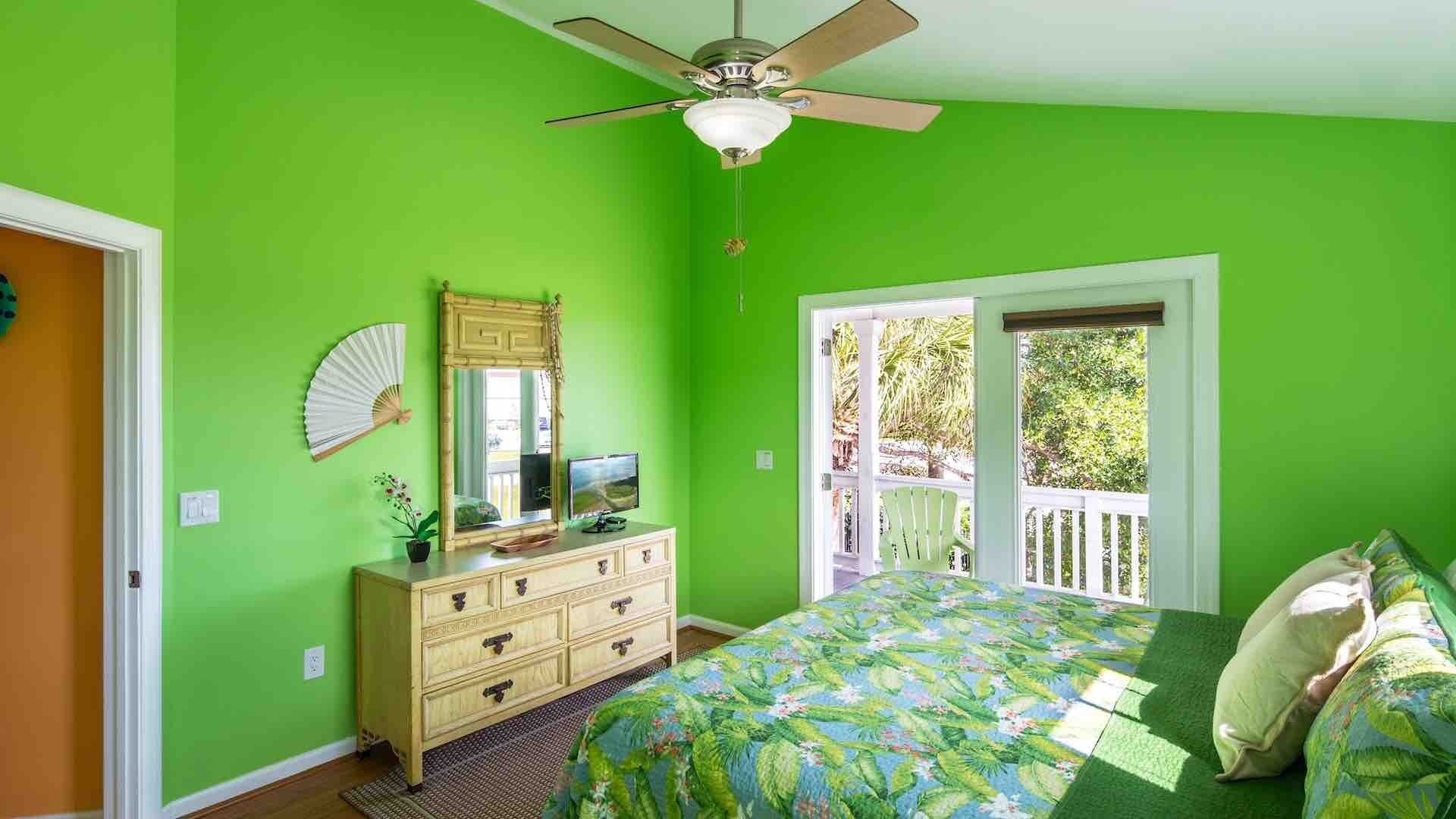 A bedroom with a green wall, a ceiling fan, a bed with a floral bedspread, a dresser, a mirror, and a window with a view of the outdoors.
