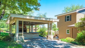 A covered patio with stone flooring, a brick fireplace, and a wooden house in the background.