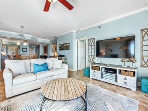 Una sala de estar con un sofá blanco, una mesa de centro de madera, un televisor y una cocina al fondo.