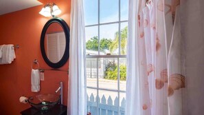 Un baño con un espejo redondo, un lavabo de cristal y una ventana que da a vistas de vegetación y una cerca.