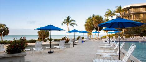 A poolside area with white lounge chairs and blue umbrellas, overlooking a body of water and palm trees.