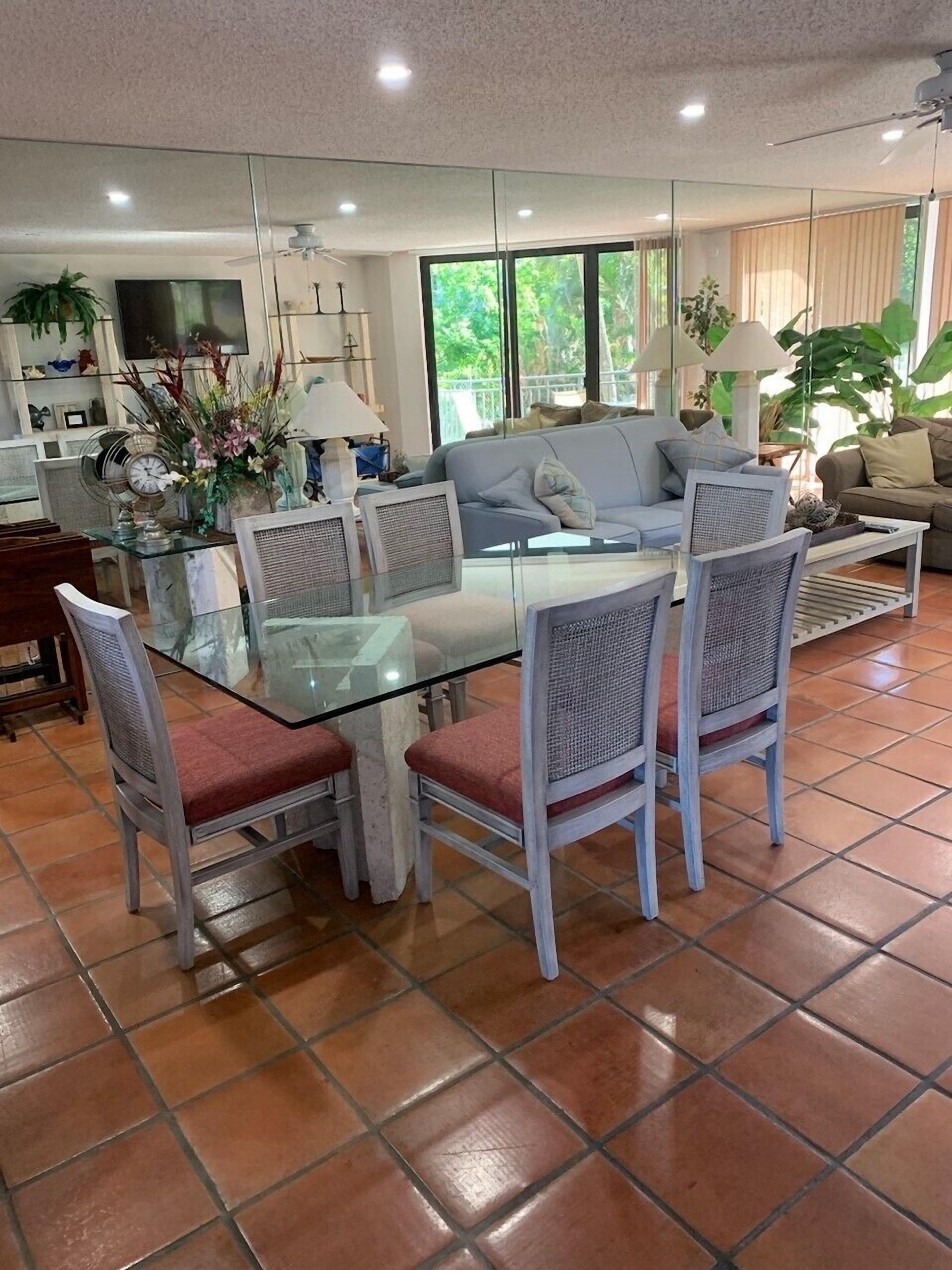 A modern living room with a glass dining table, white wicker chairs, a grey sofa, and a large window offering a view of greenery.