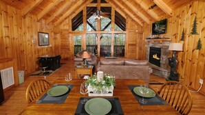 A wooden cabin interior with a dining table set for two, a fireplace, a television mounted on the wall, and a large window overlooking a forest.