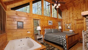 A log cabin bedroom with a large bed, a wooden headboard, a window with blinds, a small bedside table with a lamp, and a wooden floor.