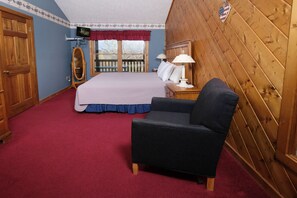 Une chambre avec un grand lit, une chaise en bois, une télévision et un balcon donnant sur la forêt.