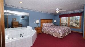 Quarto com uma banheira grande e branca, uma cabeceira de madeira, um tapete vermelho e uma janela com vista para um deck de madeira.