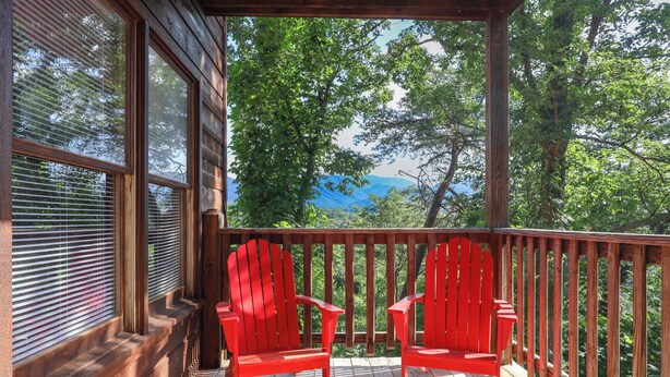 Um deck de madeira com duas cadeiras Adirondack vermelhas, corrimãos de madeira e vista para árvores e montanhas.