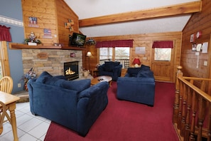 A cozy cabin interior with a stone fireplace, blue sofas, a wooden staircase, and a wooden ceiling.