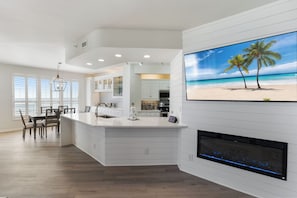 A modern kitchen with a large island, white cabinetry, and a flat-screen TV displaying a beach scene. - 904E Gulf Front unit Breathtaking Views of Gulf! (Orange Beach)