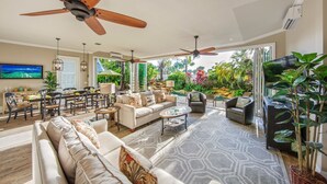 A spacious living room with a large sofa, a coffee table, and a dining area with chairs and a table. There is a ceiling fan, a TV, and a view of a lush garden through large windows.