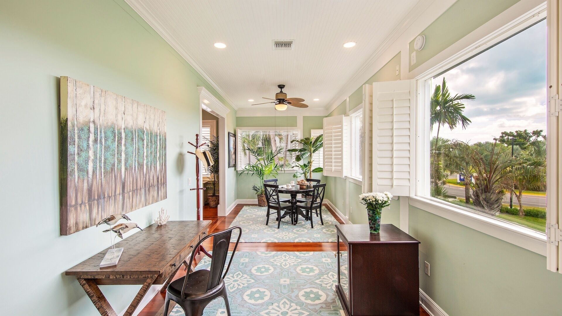 A dining area with a wooden table, chairs, and a ceiling fan. There are potted plants and a view of palm trees outside.