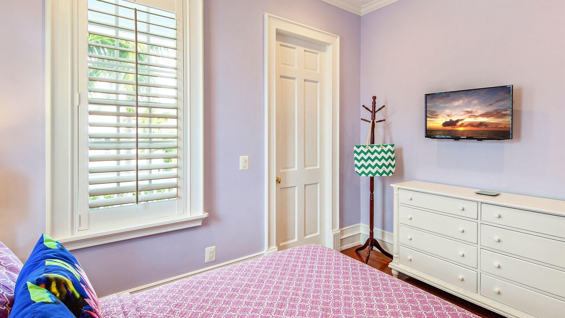 A bedroom with a bed, a dresser, a window with blinds, a door, and a wall-mounted TV displaying a sunset scene.