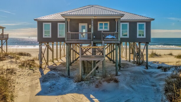 A wooden beach house with multiple decks and large windows, situated on stilts over the sand.