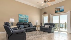 A spacious living room with two black leather sofas, a coffee table, and a ceiling fan. There are two paintings on the walls, a window with a view of the ocean, and a sliding glass door leading to a balcony.